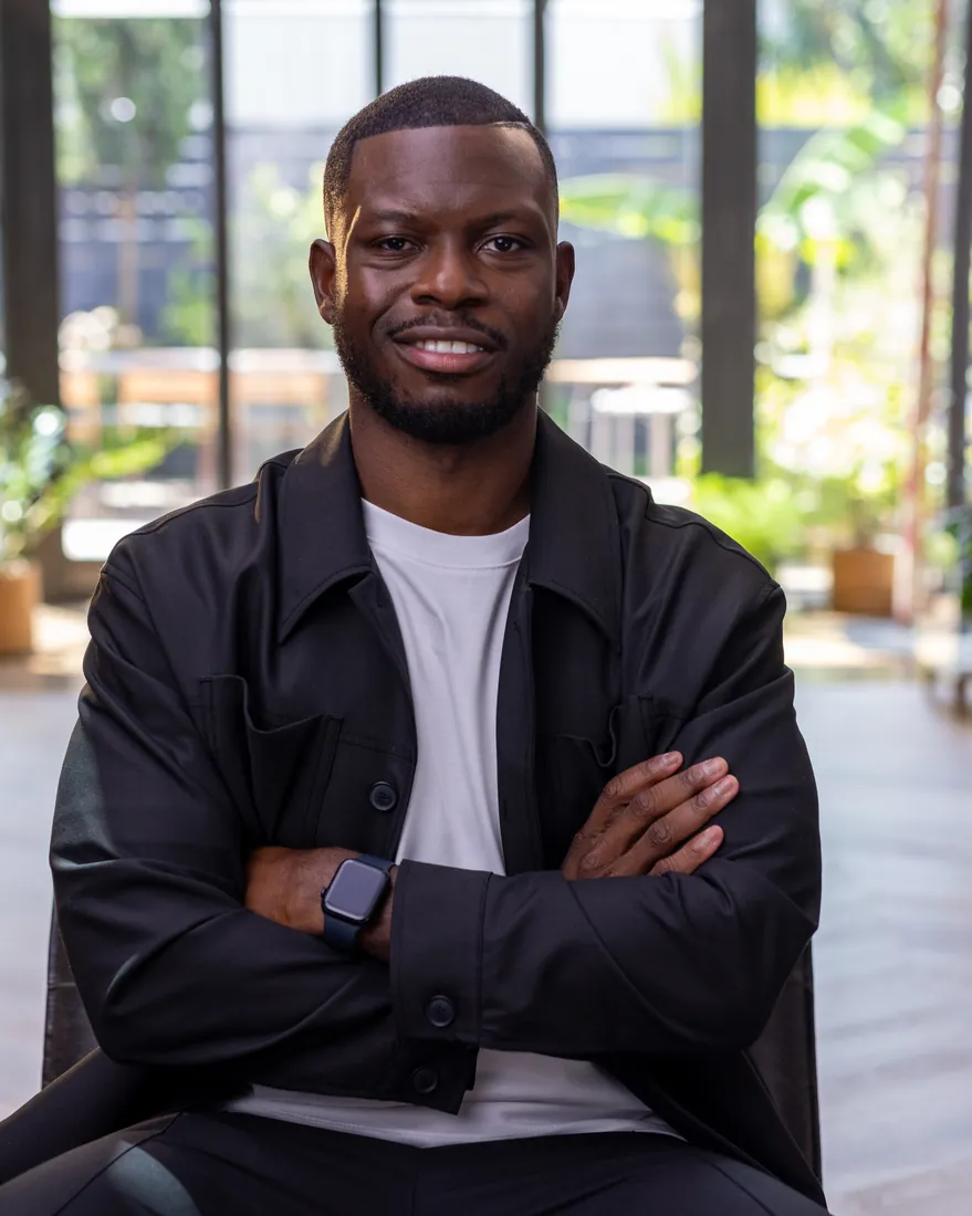Tobi Oluwole seated, arms crossed, in a bright plant-filled studio
